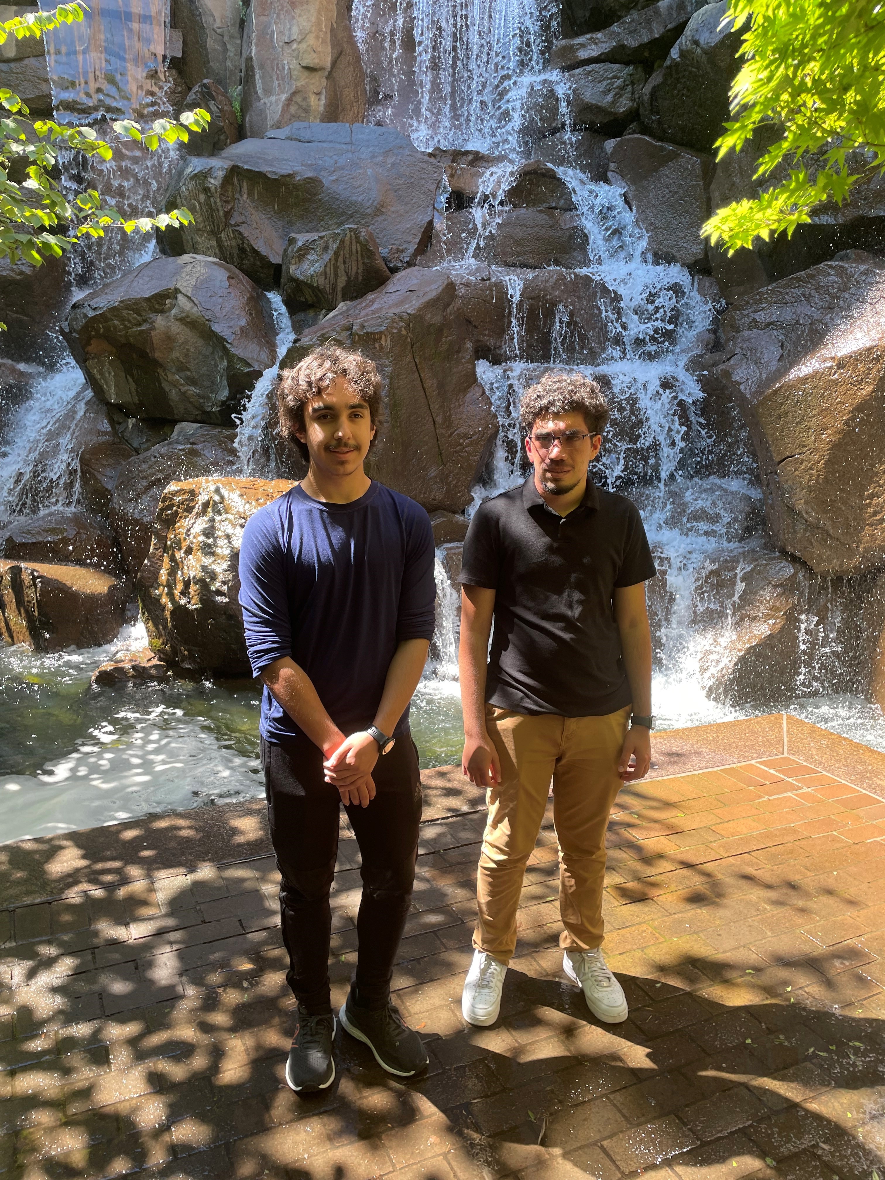 photograph of two masculine-presenting young adults standing in front of a waterfall fountain in downtown Seattle