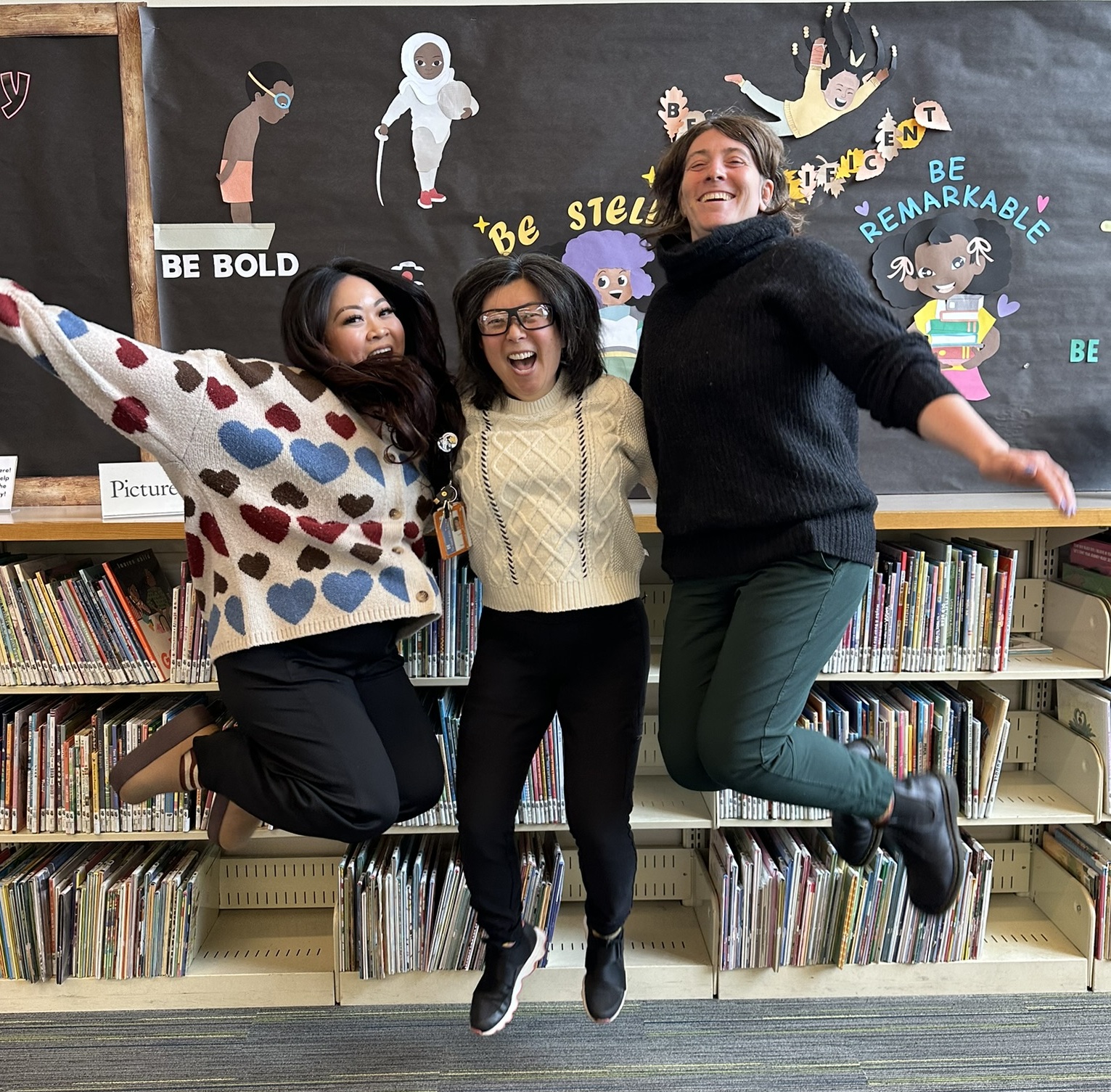 Three women jumping with expressions of joy on their face. In the background are library bookshelves and a display with illustrations and affirming messages.