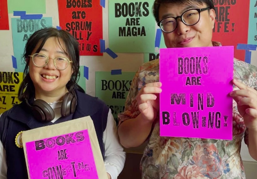Photo of two people holding letterpress posters about books, against a backdrop of more letterpress posters.